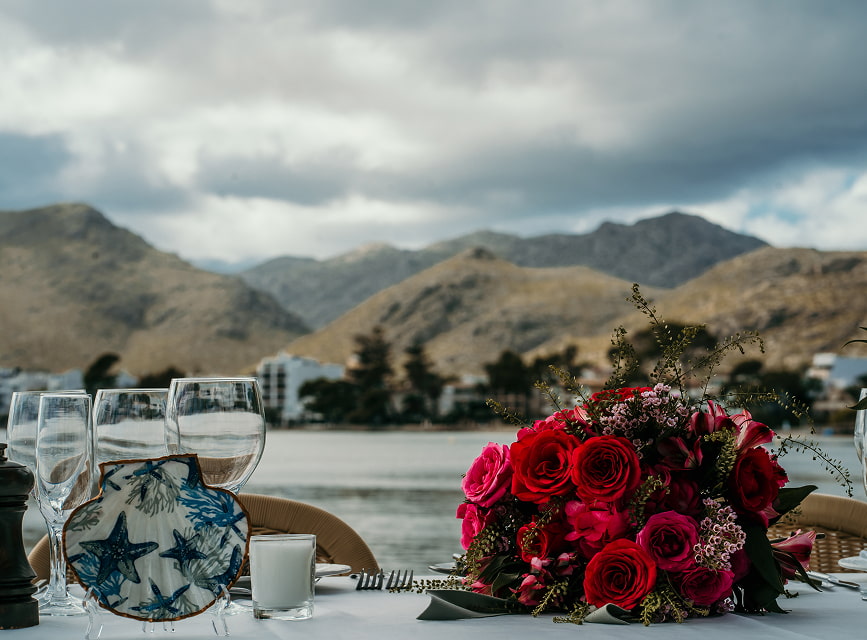 Tisch mit roten Rosen, Gläsern und blauer Keramik vor der Bucht von Pollença und ihren Bergen