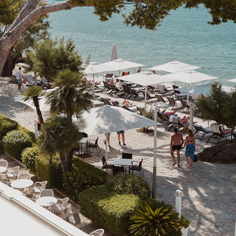 Hotelgarten mit Tischen an der Strandpromenade und Liegen mit Sonnenschirmen am Meer