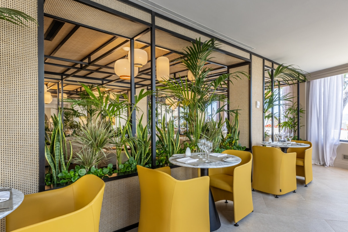 Speisesaal des Restaurants mit gelben Stühlen, runden Tischen, Pflanzen und verglasten Wänden