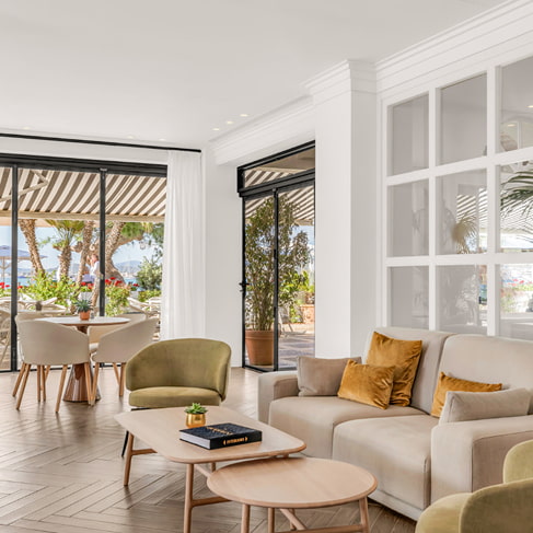 Helle Lounge mit Sofa, Sesseln und Couchtischen an großen Fenstern zur Terrasse und zum Garten