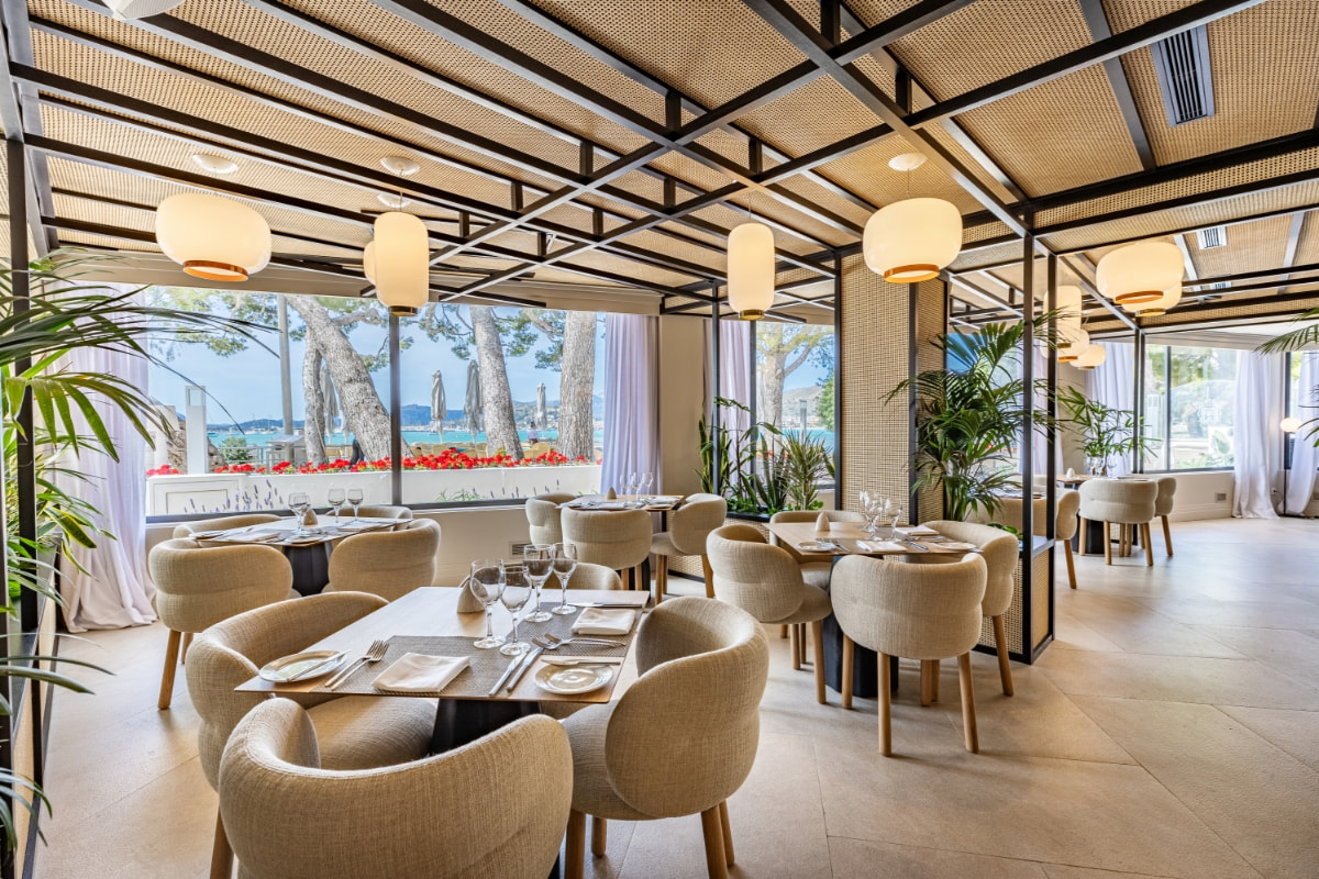 Heller Speisesaal mit gedeckten Tischen, Polsterstühlen und geflochtenen Lampen unter einer Pergola, mit Meerblick