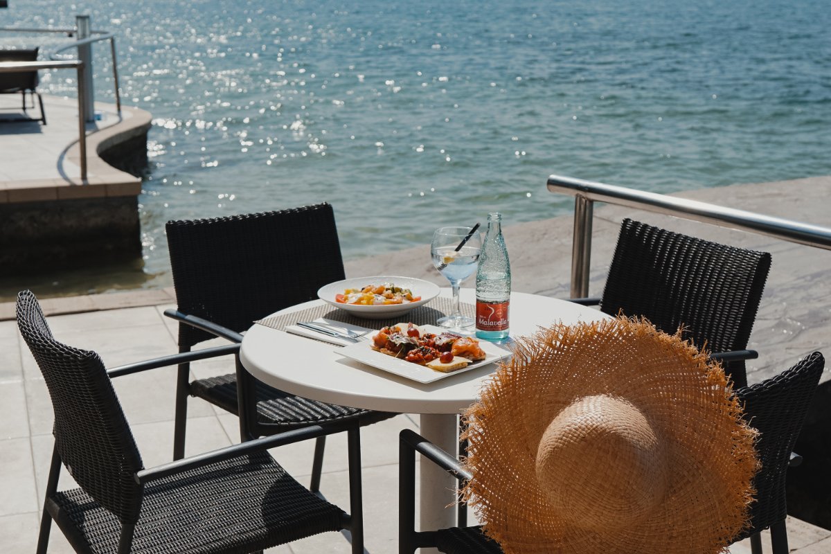 Tisch direkt am Wasser mit Speisen, Getränken und einem Strohhut auf der Terrasse an der Bucht
