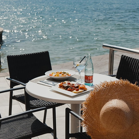 Tisch direkt am Wasser mit Speisen, Getränken und einem Strohhut auf der Terrasse an der Bucht