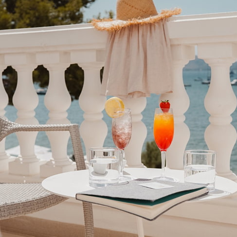 Zwei Cocktails und eine Karte auf einem Tisch auf der Terrasse mit weißer Balustrade und Blick auf die Bucht