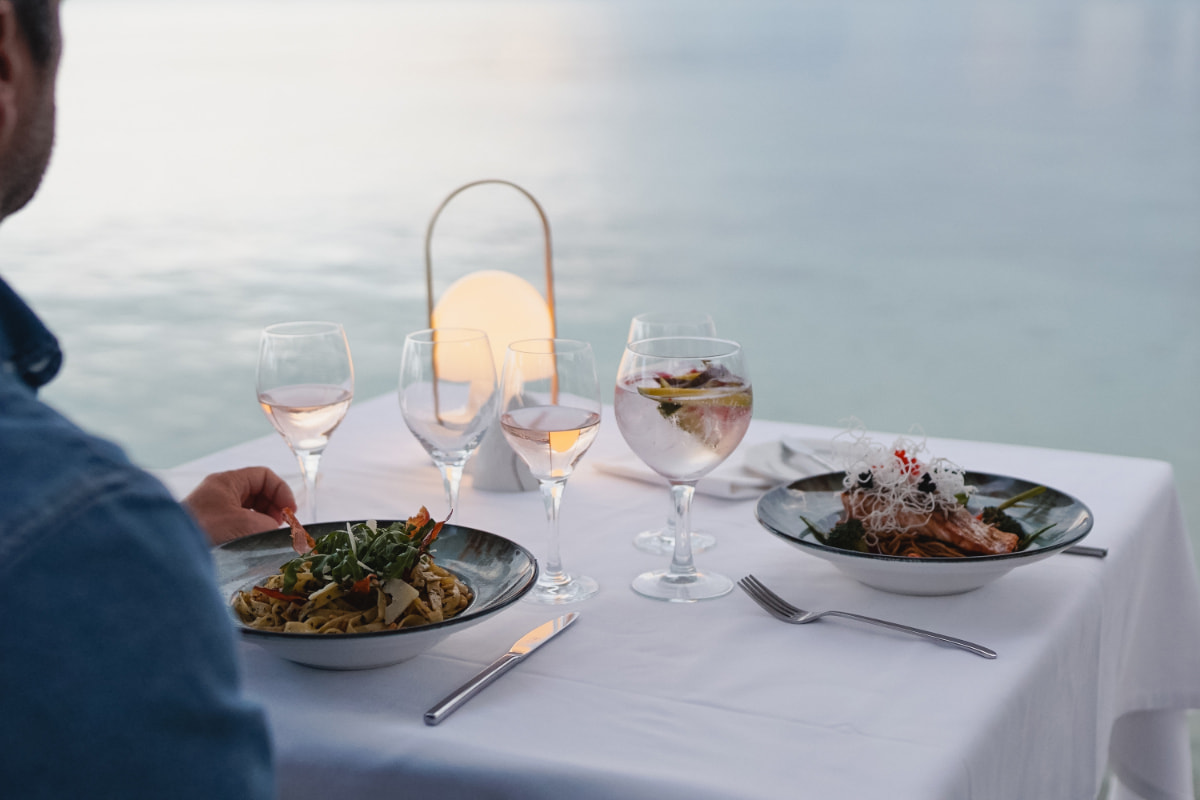 Tisch für zwei mit weißer Tischdecke, Pastagerichten, Roséwein und brennender Laterne am Meer