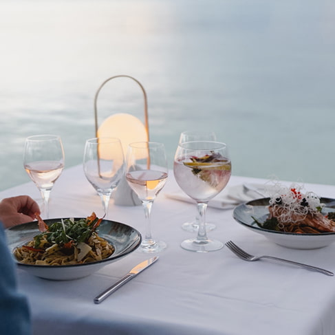 Tisch für zwei mit weißer Tischdecke, Pastagerichten, Roséwein und brennender Laterne am Meer