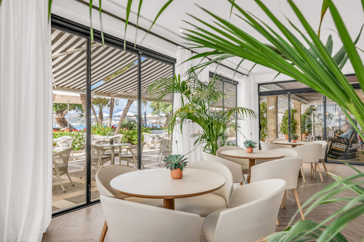 Café-Bereich mit runden Tischen, weißen Sesseln und Pflanzen an großen Fenstern zur Terrasse