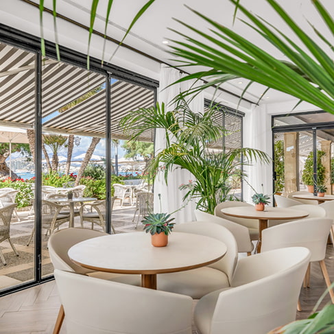 Café-Bereich mit runden Tischen, weißen Sesseln und Pflanzen an großen Fenstern zur Terrasse