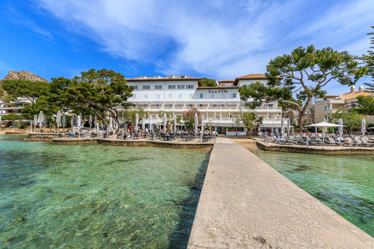 Weiße Fassade des Hotel Illa d'Or am Meer, vom Steg über kristallklares Wasser in Puerto Pollensa gesehen