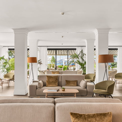 Großzügige Lounge mit Sofas, Sesseln, Stehlampen und weißen Säulen, durch große Fenster erhellt
