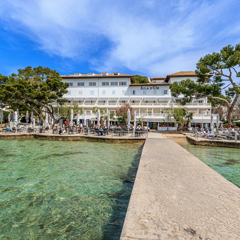 Weiße Fassade des Hotel Illa d'Or am Meer, vom Steg über kristallklares Wasser in Puerto Pollensa gesehen