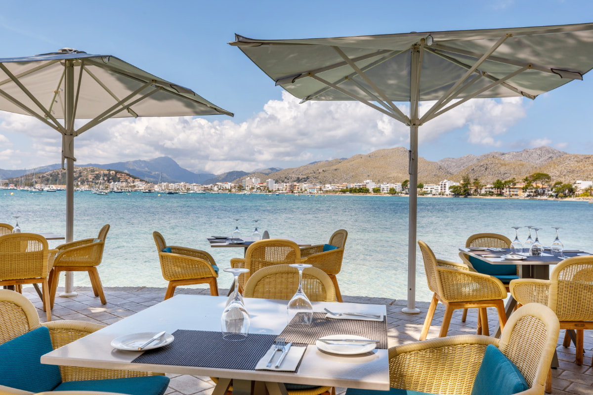 Gedeckte Tische auf der Restaurantterrasse unter Sonnenschirmen mit Blick auf Bucht und Berge von Pollença