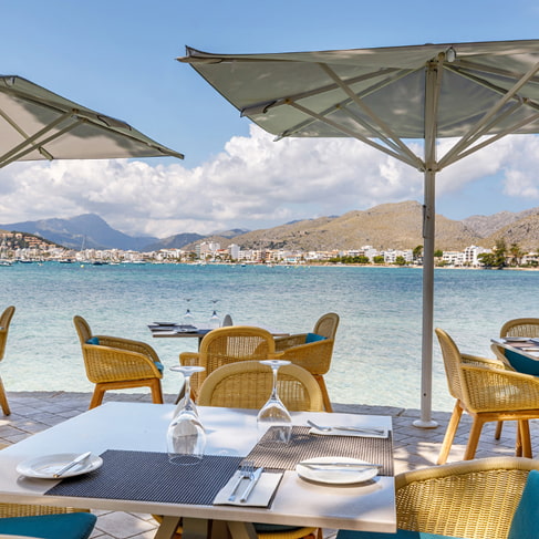 Gedeckte Tische auf der Restaurantterrasse unter Sonnenschirmen mit Blick auf Bucht und Berge von Pollença