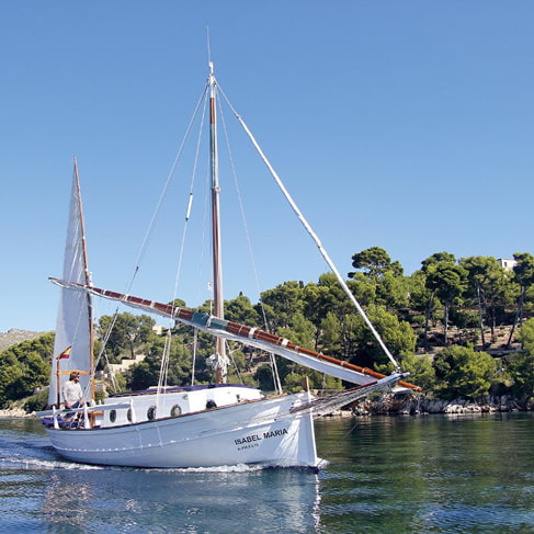 Traditionelles mallorquinisches Llaüt-Segelboot entlang der Küste, im Hintergrund Pinienwälder