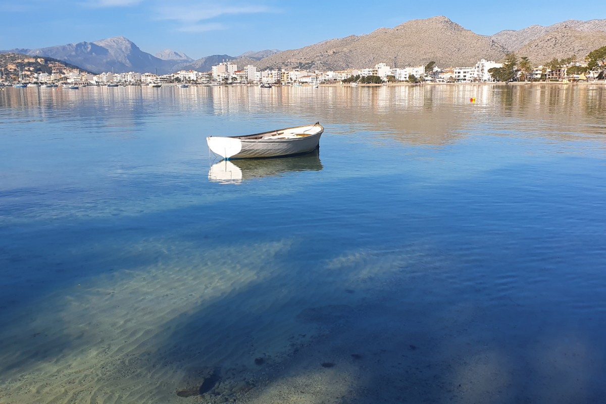 Kleines weißes Boot in kristallklarem Wasser, dahinter die Uferfront von Puerto Pollensa und die Berge