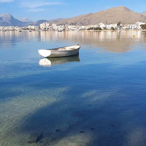 Kleines weißes Boot in kristallklarem Wasser, dahinter die Uferfront von Puerto Pollensa und die Berge
