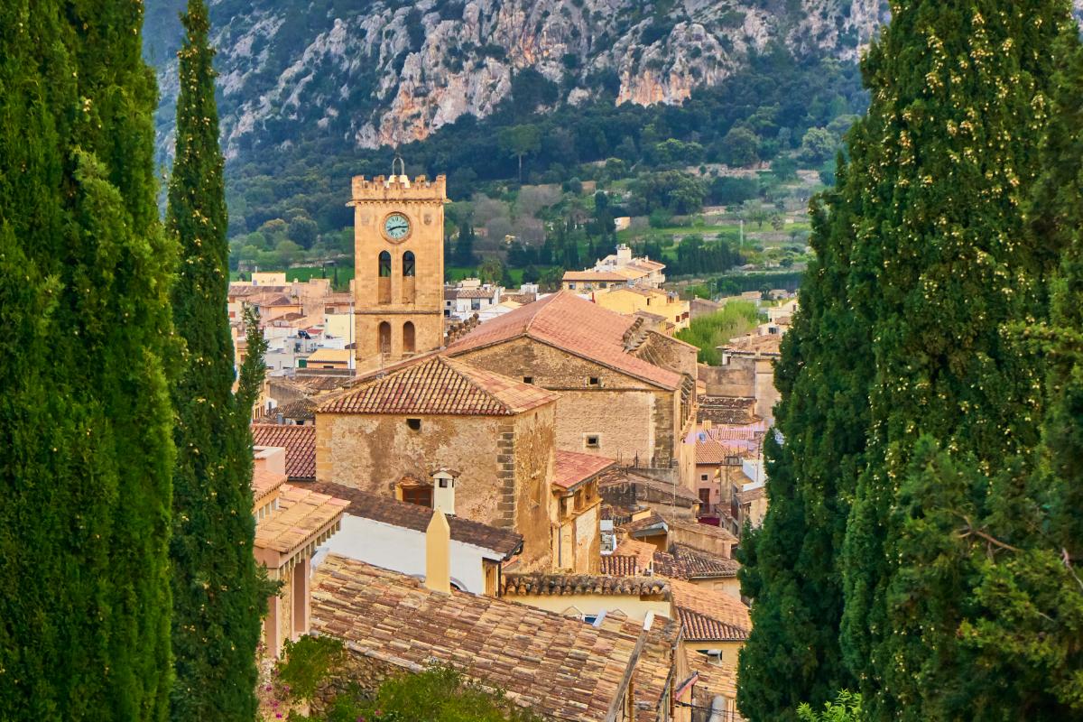 Dächer und Kirchturm der Altstadt von Pollença zwischen Zypressen, dahinter die Serra de Tramuntana