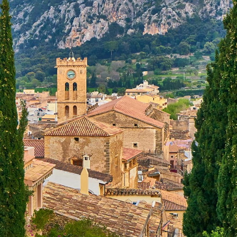 Dächer und Kirchturm der Altstadt von Pollença zwischen Zypressen, dahinter die Serra de Tramuntana