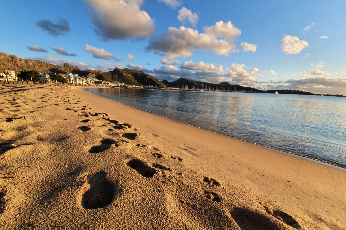 Fußspuren im Sand am Strand von Puerto Pollensa bei Sonnenuntergang, im Hintergrund Küste und Berge