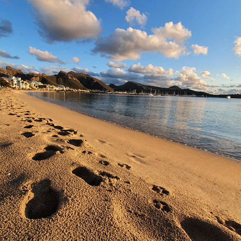 Fußspuren im Sand am Strand von Puerto Pollensa bei Sonnenuntergang, im Hintergrund Küste und Berge
