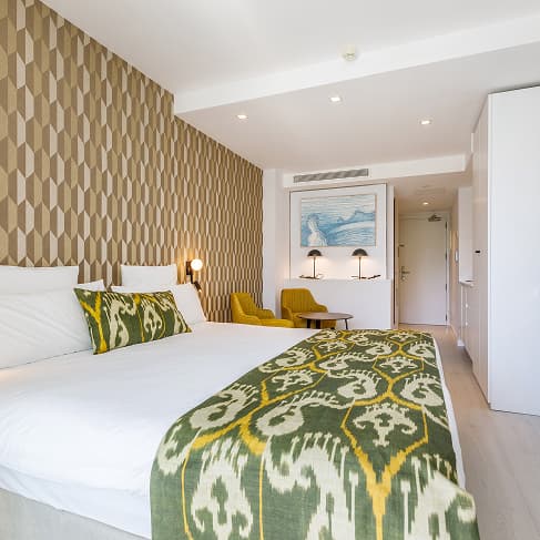 Doppelzimmer mit geometrischer Tapete, mallorquinischer Tagesdecke sowie Theke mit Hockern und Kaffeemaschine