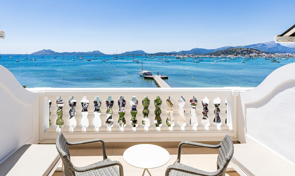 Balkon mit zwei Stühlen und Tisch direkt an der Bucht von Pollença, im Hintergrund Steg und Berge