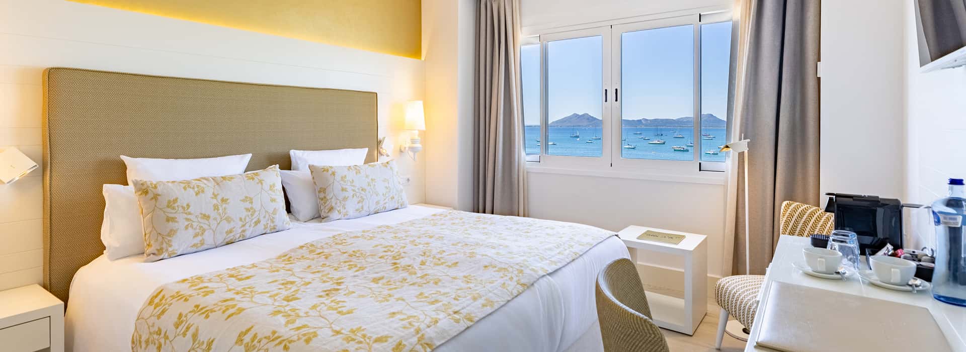 Hotel Illa d'Or | Sea View Room Room with a double bed featuring a floral bedspread in golden tones and a window overlooking the sea