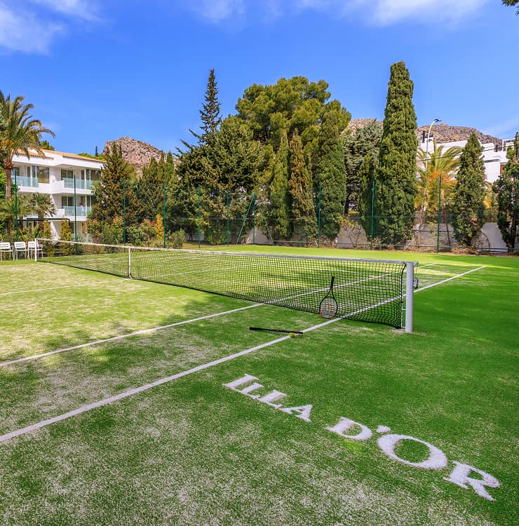 Pista de tenis de hierba con el logotipo Illa d'Or, rodeada de pinos y cipreses junto al hotel