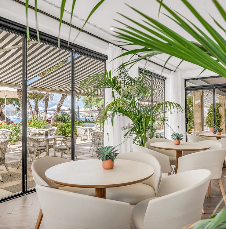 Espacio luminoso con mesas redondas, sillas tapizadas y plantas junto a grandes ventanales