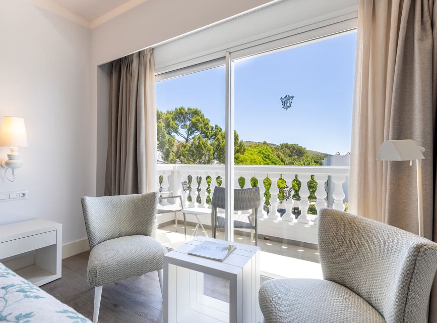 Armchairs and side table beside the large window opening onto a balcony with white balustrade and greenery