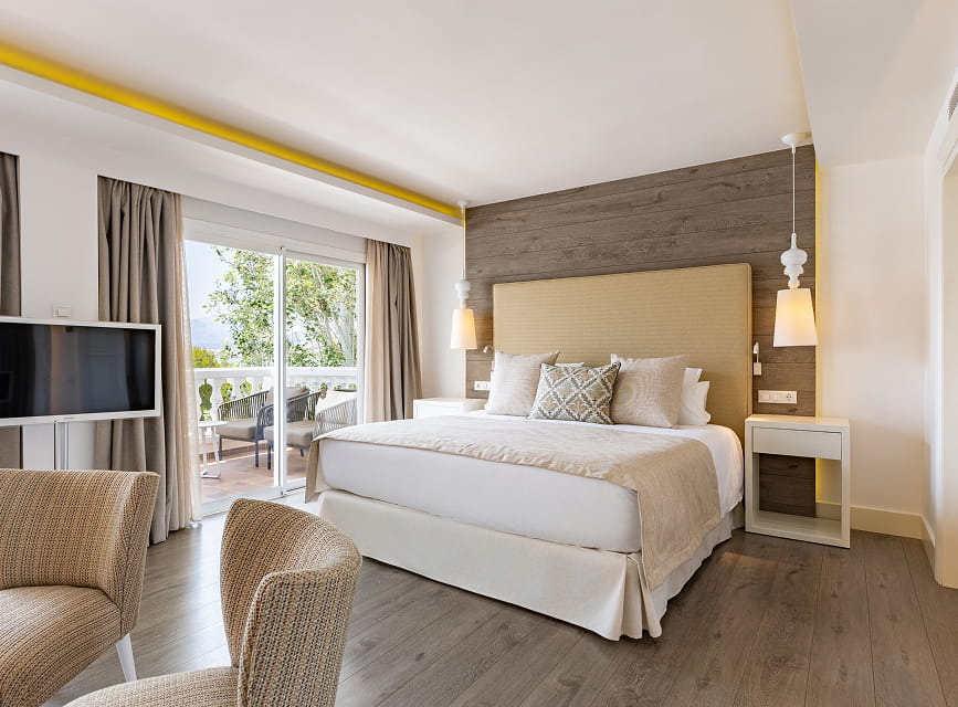 Suite bedroom with double bed, upholstered headboard, wood-panelled wall, armchairs and terrace access