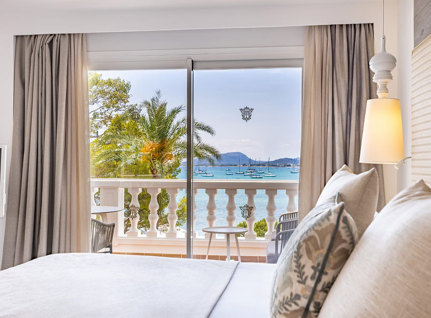 Suite bed facing the large window onto the terrace, with palm trees and Pollensa Bay beyond