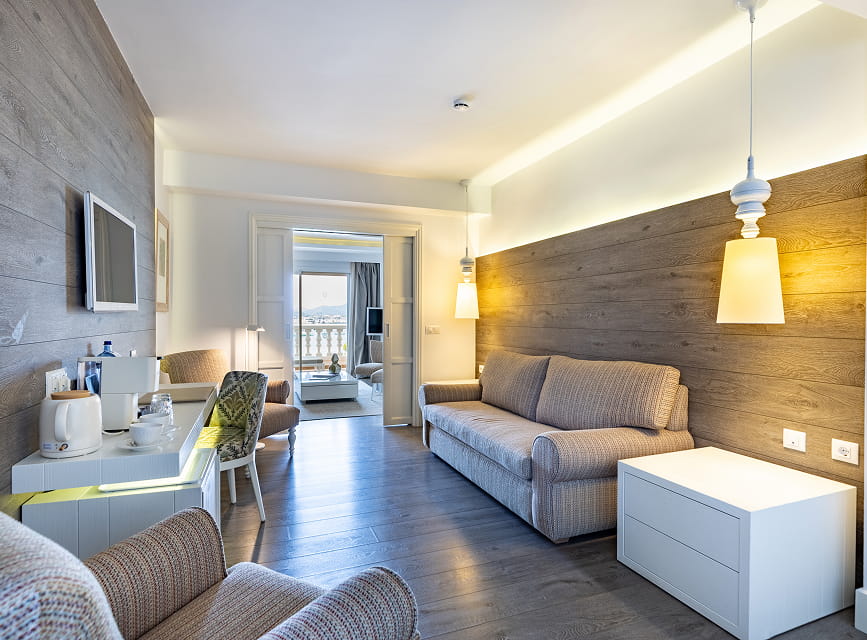 Suite living room with sofa, wood-panelled wall, minibar and coffee machine, open to the bedroom
