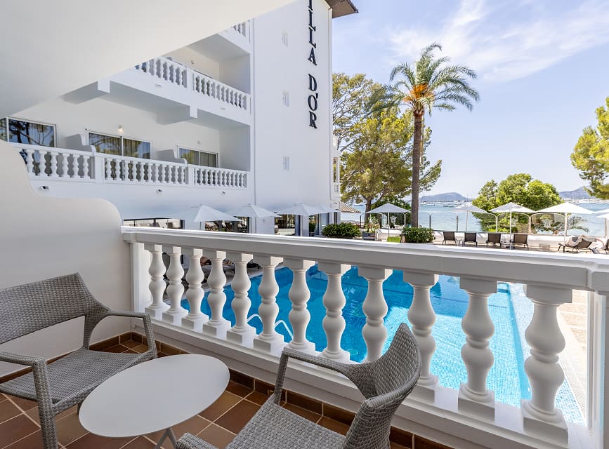 Balcony with chairs and table above the hotel pool, with palm trees and the sea of Puerto Pollensa behind